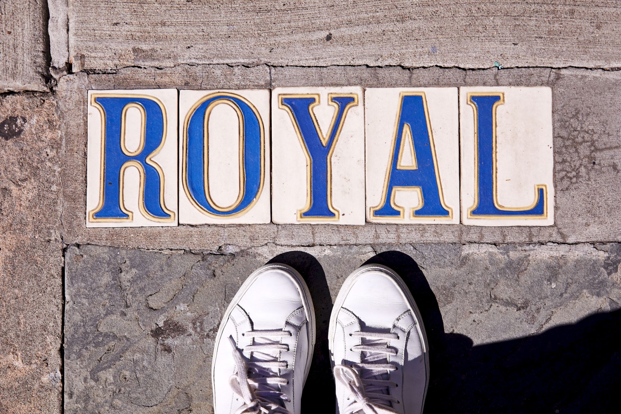 The image shows white sneakers on a sidewalk near tiles spelling "ROYAL" in blue and yellow letters.