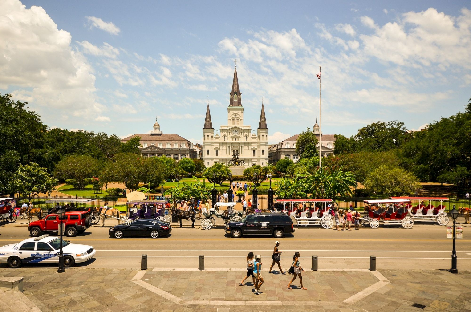 A historic building with spires, cars, people, and horse-drawn carriages in front of a park on a sunny day.