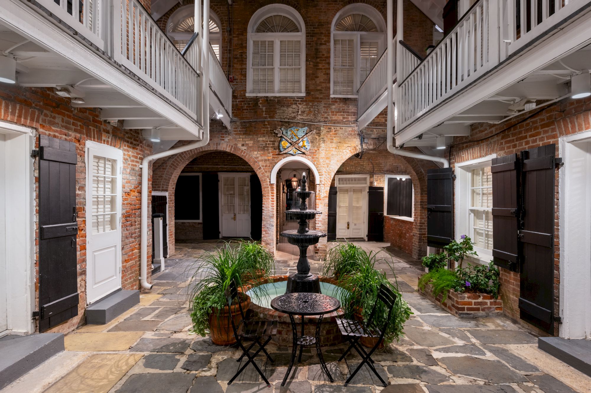 A courtyard with a central fountain, brick walls, arched doorways, and overhead walkways, surrounded by plants and chairs at the center.