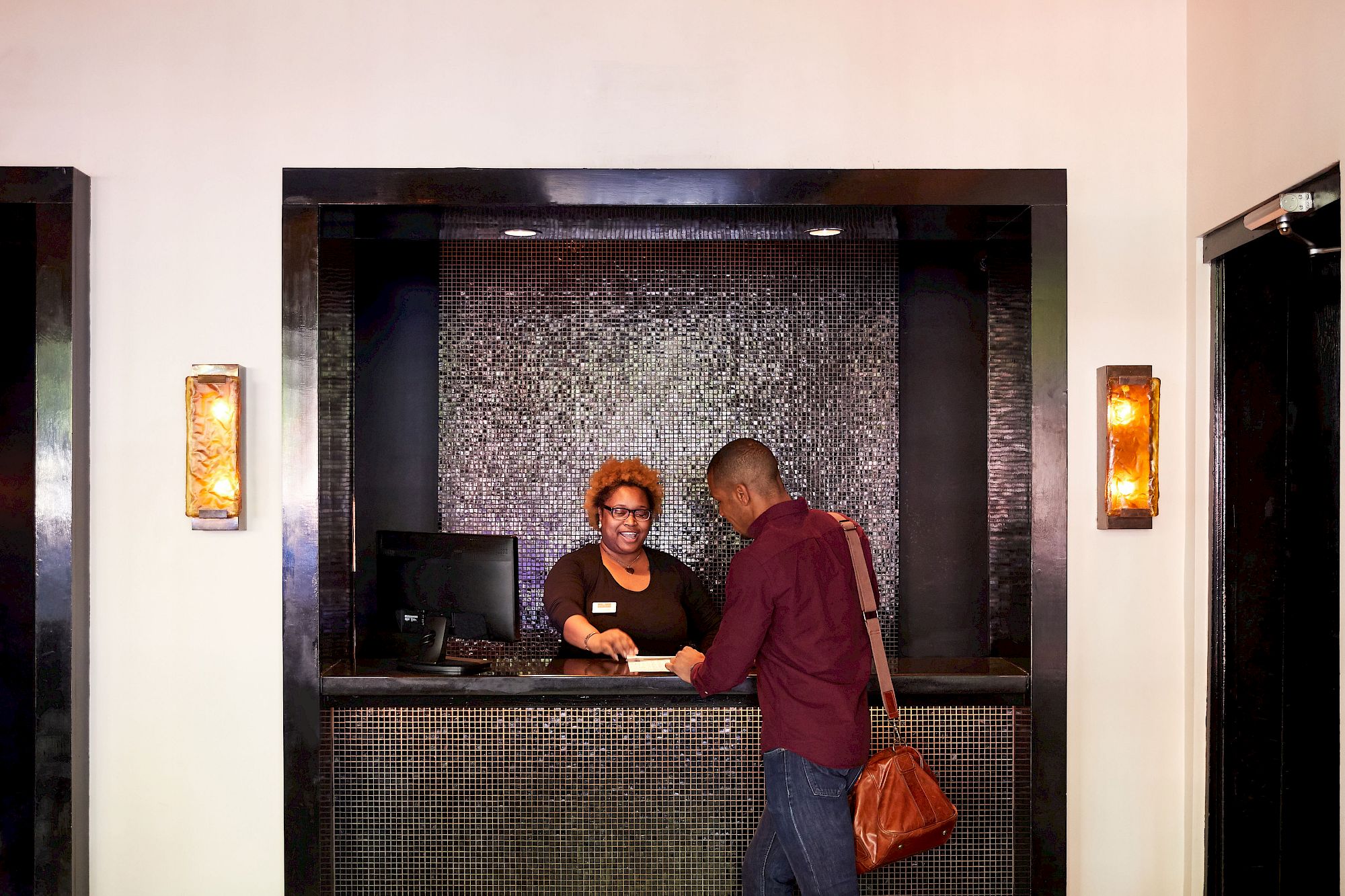 A man is interacting with a receptionist at a counter. The reception area has dark decor and soft lighting.