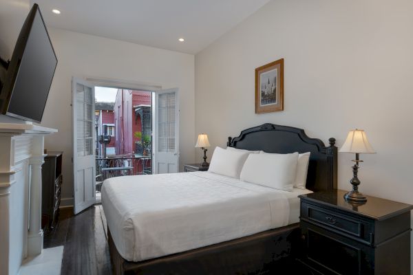 A cozy bedroom with a large bed, two lamps on side tables, a TV, and a window door opening to a balcony with a view outside.