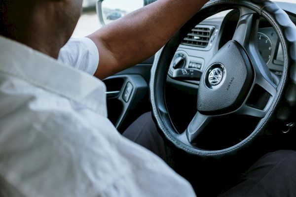 A person in a white shirt is driving a car, holding the steering wheel with one hand.