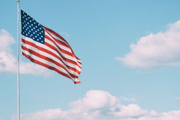 A U.S. flag waves against a clear blue sky with a few clouds visible in the background.