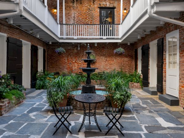 A charming courtyard with a central fountain, surrounded by plants, white railings, brick walls, and seating for two on stone flooring.