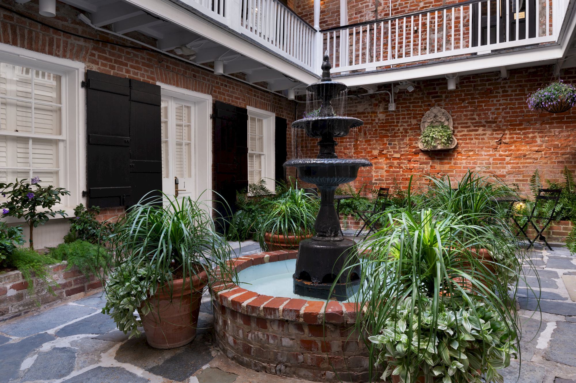 A courtyard with a central fountain is surrounded by lush potted plants, brick walls, and balconies, creating a serene atmosphere.