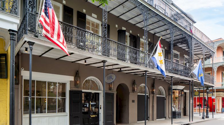 A historic building with wrought iron balconies, displaying three flags, stands on a quiet street, exuding Southern architectural charm.