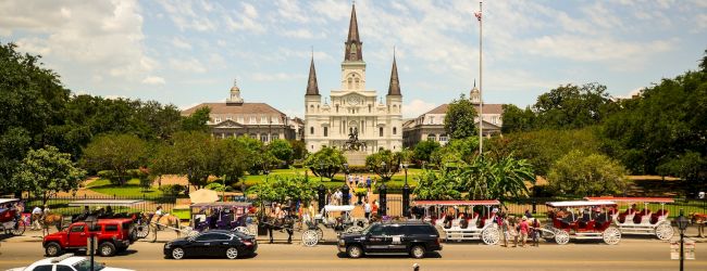 The image shows a historic building with spires, people walking, vehicles, and a park with lush greenery under a partly cloudy sky.