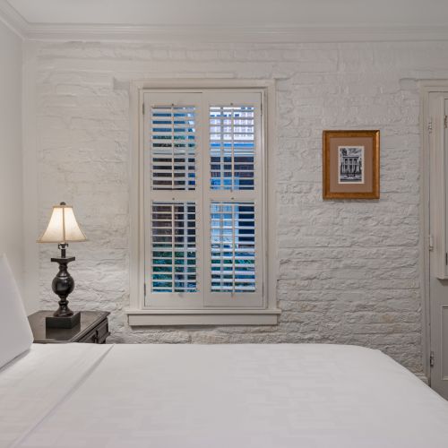 A cozy bedroom with a white bed, lamp on a bedside table, brick wall, shuttered windows, and framed picture.