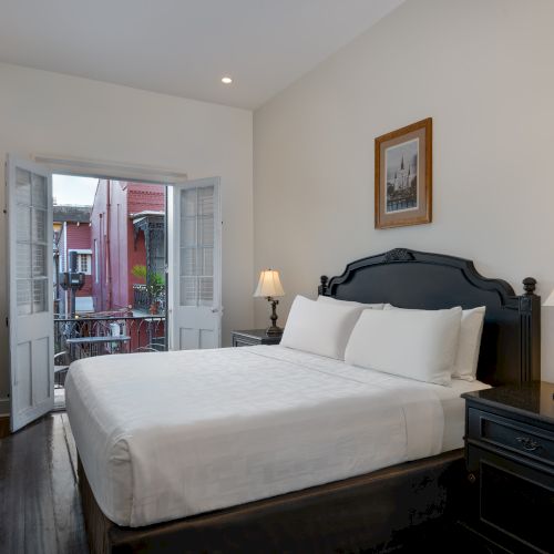 A cozy bedroom features a large bed, bedside tables with lamps, a wall-mounted TV, and an open balcony door revealing a view outside.