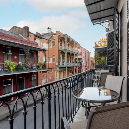 A balcony with chairs and a table overlooks a vibrant street with colorful buildings and decorative ironwork.