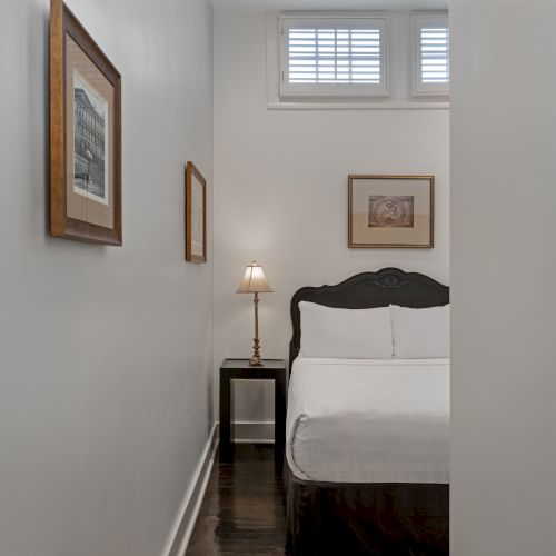 A small bedroom with a bed, side table with lamp, wall art, and window above the bed. The room is minimalist and neatly arranged.