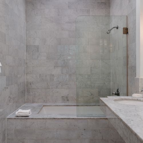 A modern bathroom features a marble-tiled shower and bathtub, dual sinks, a large mirror, and towels hanging on the wall.