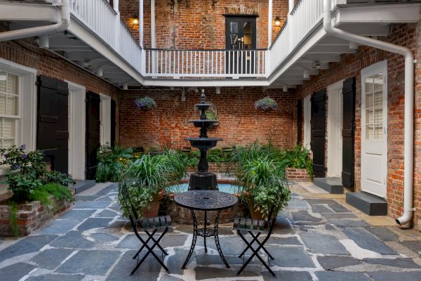 A courtyard with a central fountain, surrounded by brick walls, lush plants, and a small table with chairs on a stone floor.